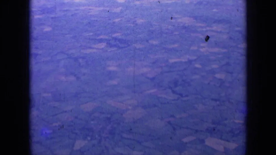 Aged 8mm aerial frame near London, 1963, with patchwork farmland, window framing, and dust specks on the scan.