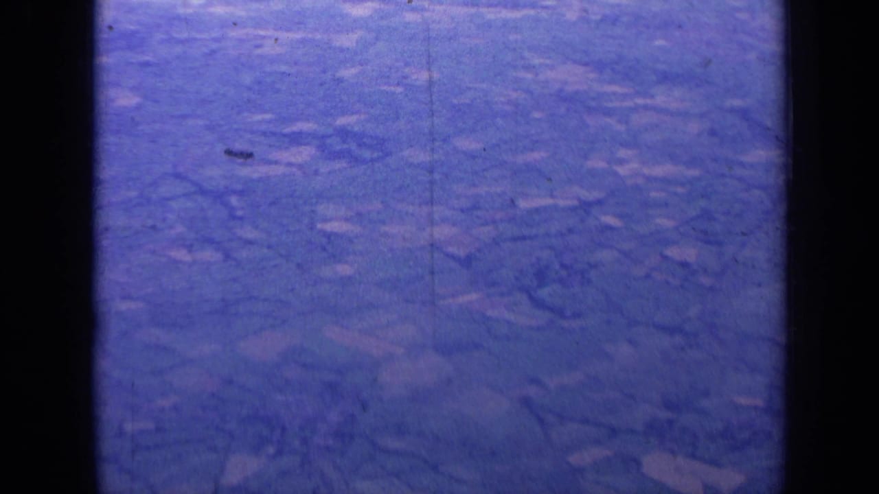 1963 8mm aerial view over rural England, showing patchwork fields and hedgerows through a blue-purple aged film cast.