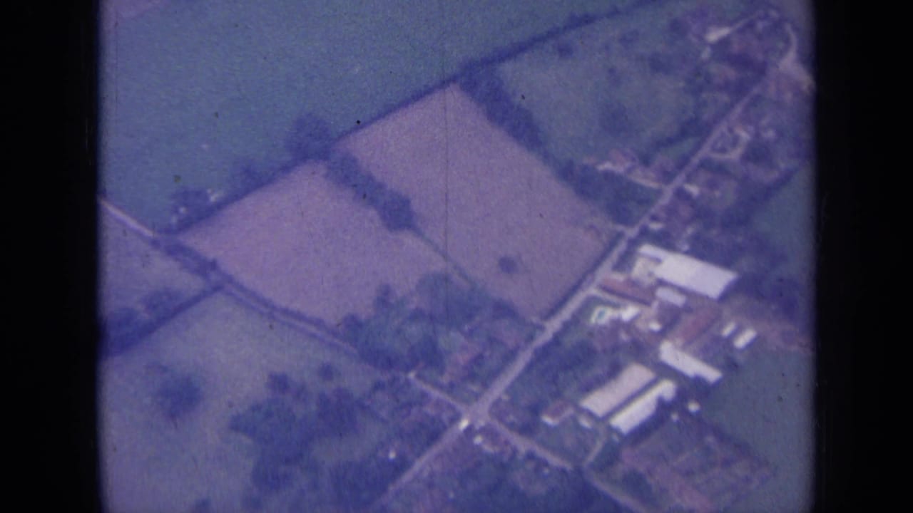 1963 aerial home-movie view over rural England, with farm buildings, lanes, and adjoining fields visible below.