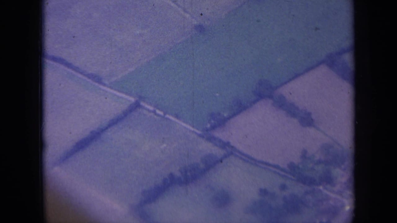 Soft 8mm aerial frame over rural England, showing rectangular fields, hedgerows, and a winding lane near the frame edge.