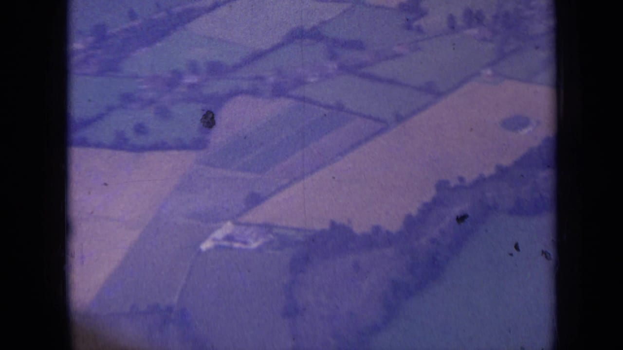 1963 8mm aerial view near London showing patchwork fields, trees, a narrow road and small rural buildings below.