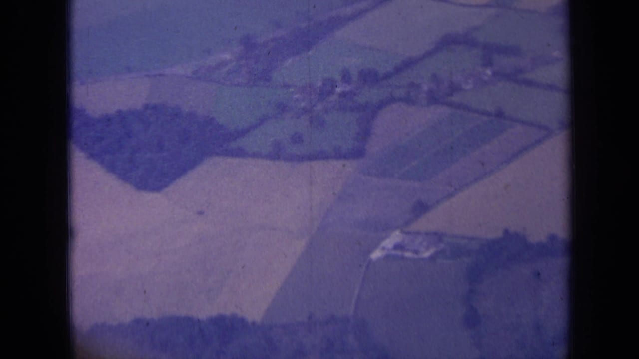 Closing 1963 London-area aerial frame with divided fields, tree clusters and pale farm structures seen through aged 8mm film.