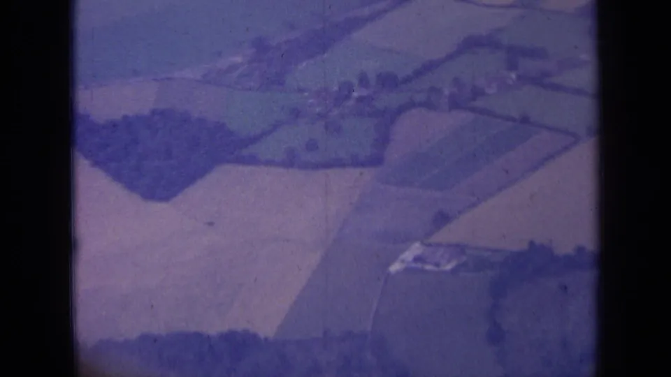 Closing 1963 London-area aerial frame with divided fields, tree clusters and pale farm structures seen through aged 8mm film.