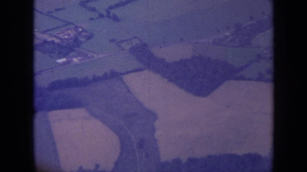 Aerial 8mm frame of rural English fields near London, showing hedgerows, lanes, and a small cluster of buildings.