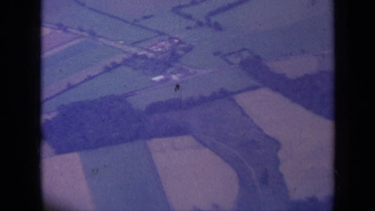 1963 aerial home-movie frame over countryside near London, with geometric fields, wooded strips, and a dark film or window divider.
