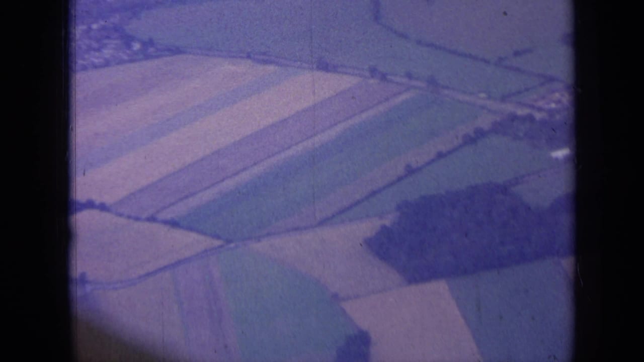 1963 aerial 8mm view over English agricultural fields with hedgerows and a dark tree cluster.