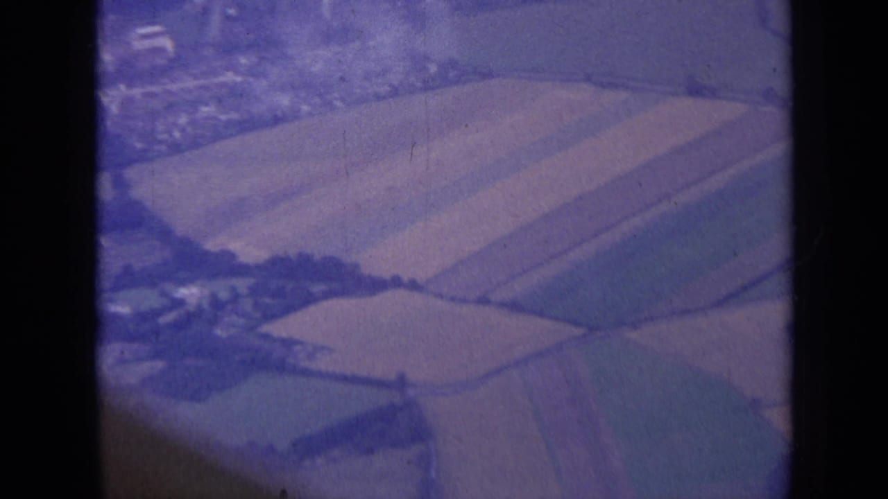 Aerial 8mm frame showing fields, hedgerows, and a nearby town edge in London, England metadata context.