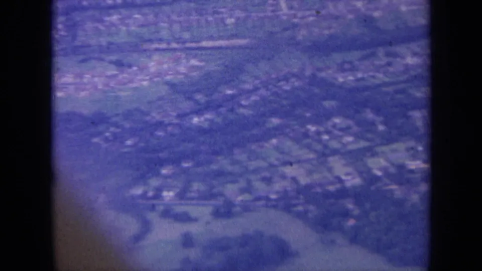 1963 aerial home-movie view over a built-up town area with rooftops, roads, and nearby fields.