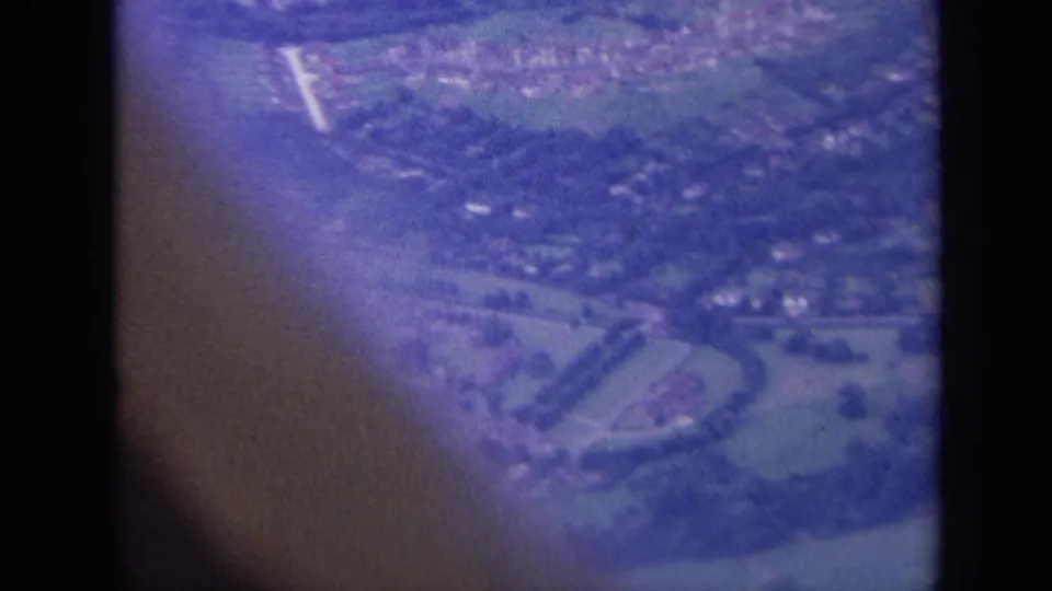 Aerial 8mm frame of a London-area town edge, with housing, roads, and fields under strong color shift.