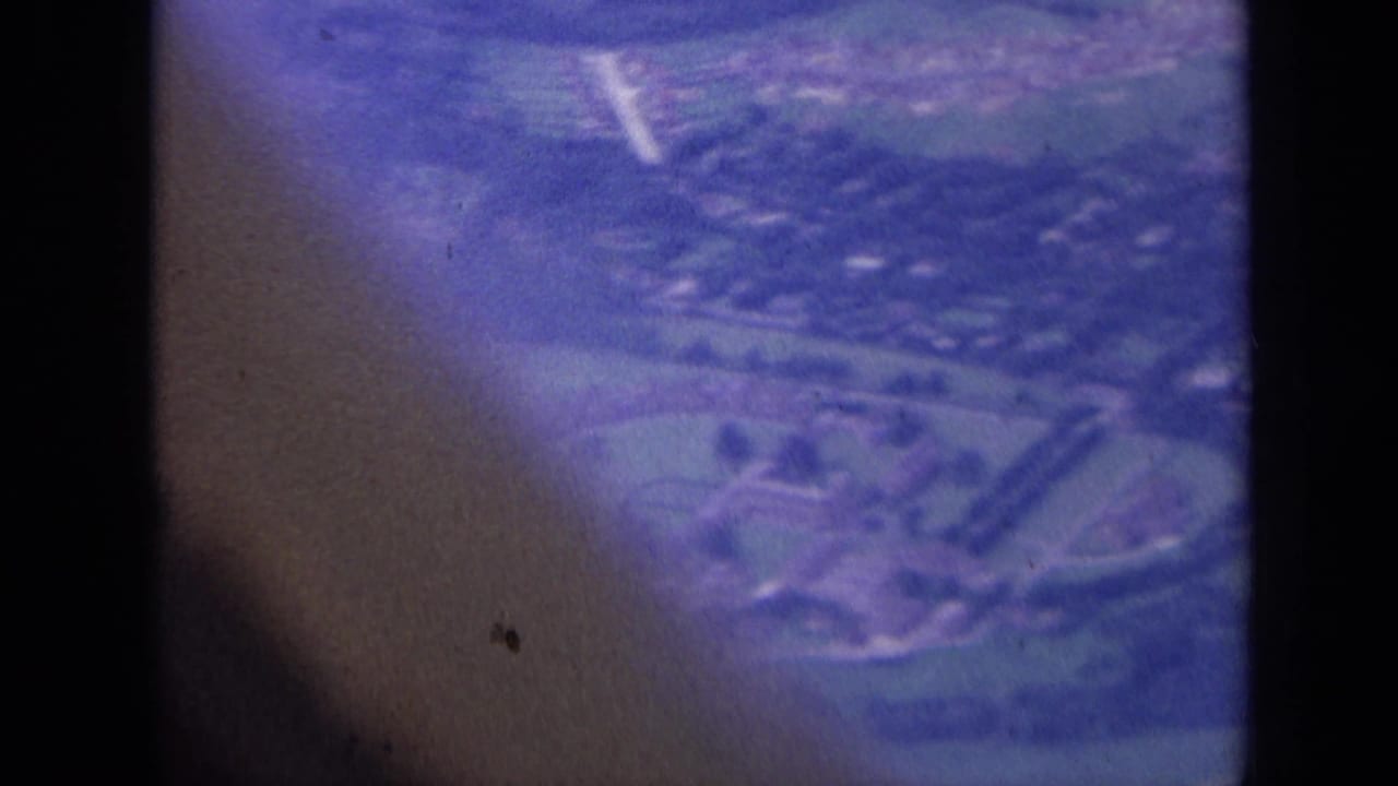 1963 London-area 8mm aerial view of a green landscape with houses and roads, softened by blue-purple film color and dark obstructions.