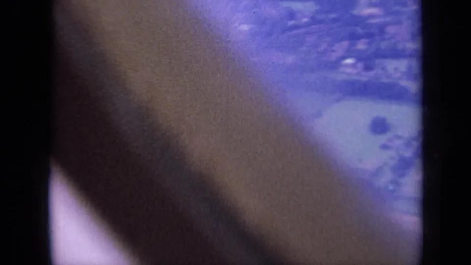 Aerial 8mm frame from the London-area clip, with a pale green field, a small dark tree or shrub, and housing softened by film aging.