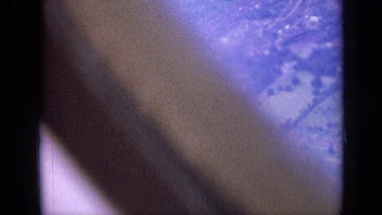 1963 8mm airplane view over London suburbs, partly blocked by a dark aircraft or window edge with hazy ground below.