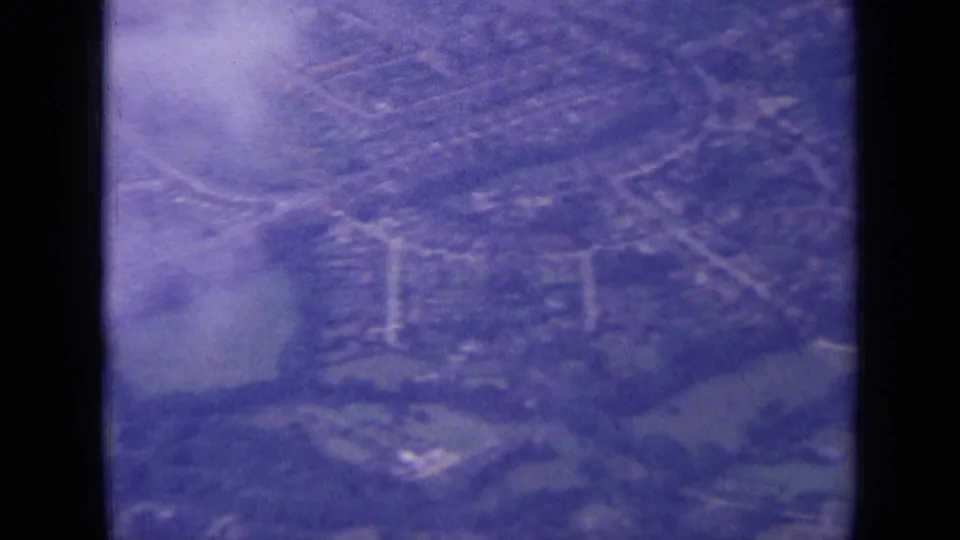 Purple-tinted airplane-window view over London suburbs, showing roads, housing, trees, and hazy open ground below.