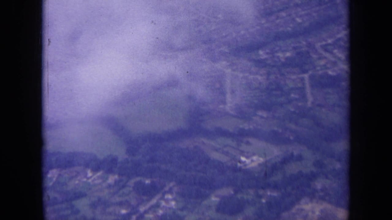 1963 8mm aerial view over the London area, with houses, roads, fields, trees, haze, and dark window edges visible.