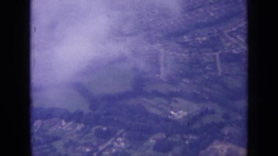 1963 8mm aerial view over the London area, with houses, roads, fields, trees, haze, and dark window edges visible.