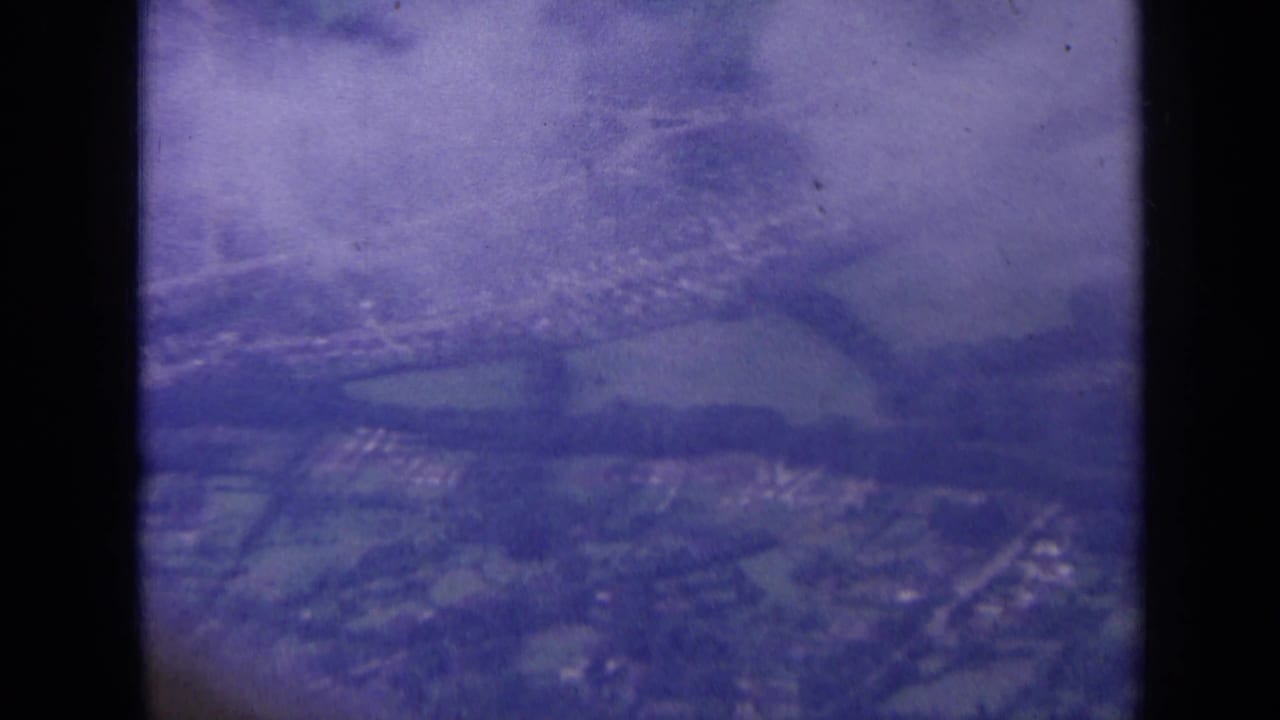 1963 London-area aerial frame showing suburban houses, roads, fields, and cloud-softened distance through the aircraft window.