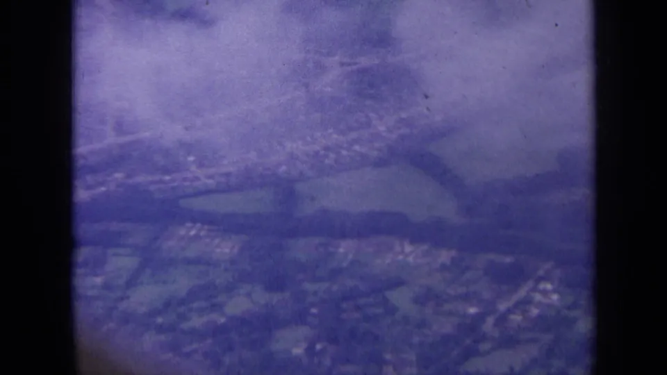 1963 London-area aerial frame showing suburban houses, roads, fields, and cloud-softened distance through the aircraft window.