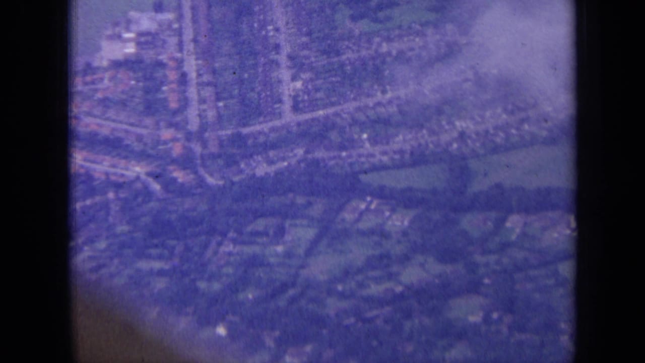 1963 8mm aerial view over a London-area residential district, with compact houses, roads, trees, and green spaces visible.