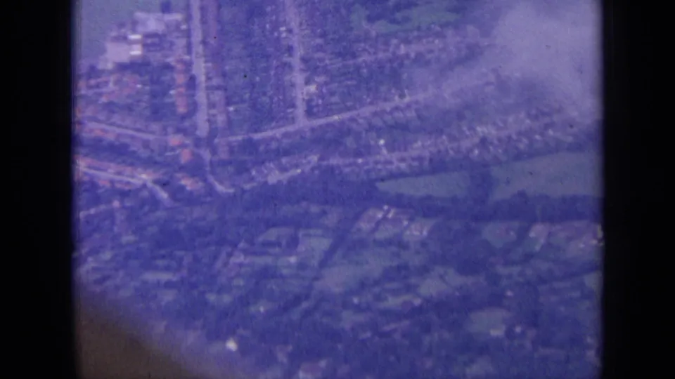 1963 8mm aerial view over a London-area residential district, with compact houses, roads, trees, and green spaces visible.