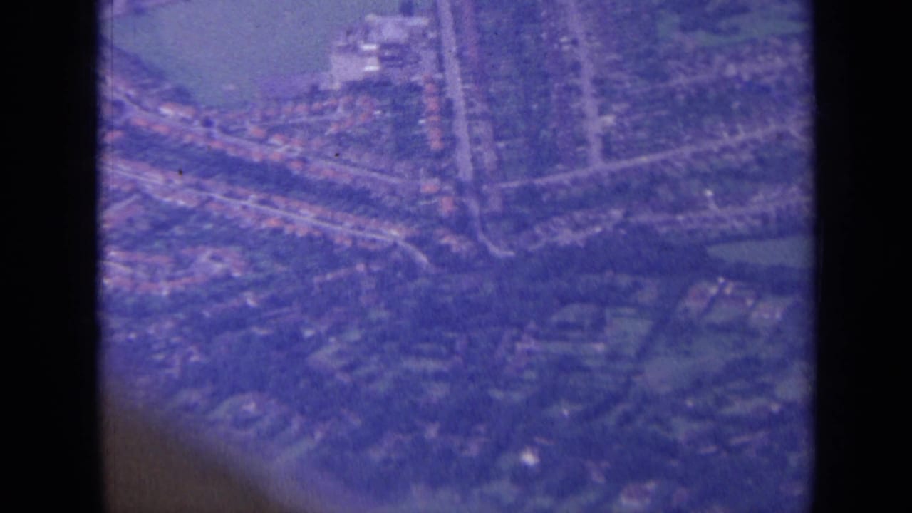 1963 London-area aerial frame showing rows of houses, a main road, trees, and a pale green or blue patch in soft 8mm color.