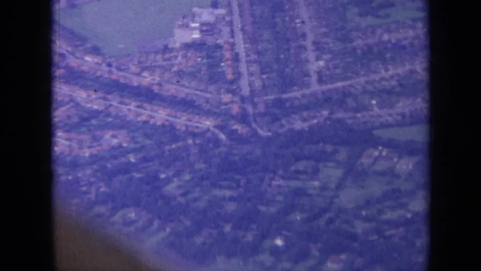 1963 London-area aerial frame showing rows of houses, a main road, trees, and a pale green or blue patch in soft 8mm color.