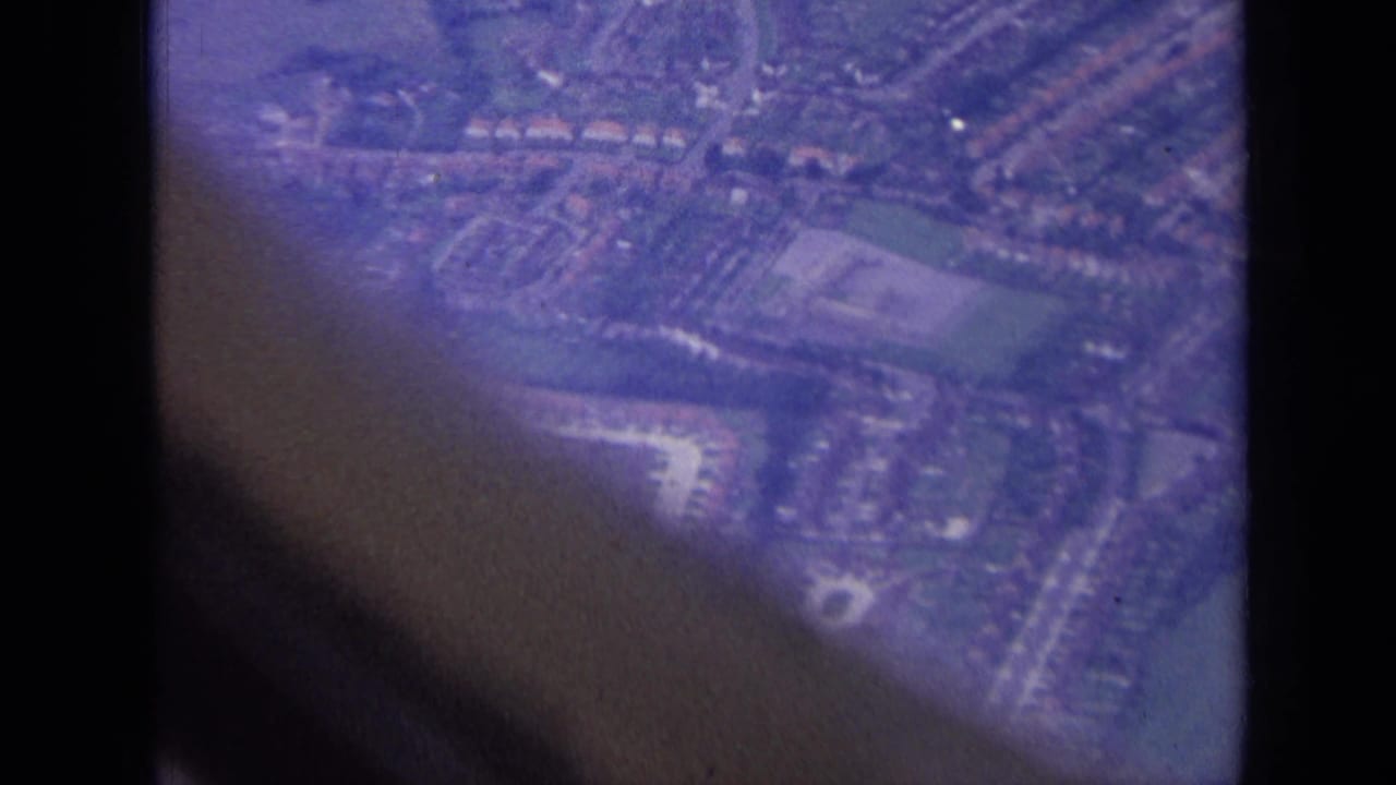 1963 aerial home-movie frame over London suburbs, with residential blocks, fields, roads, and dark aircraft-window obstruction.