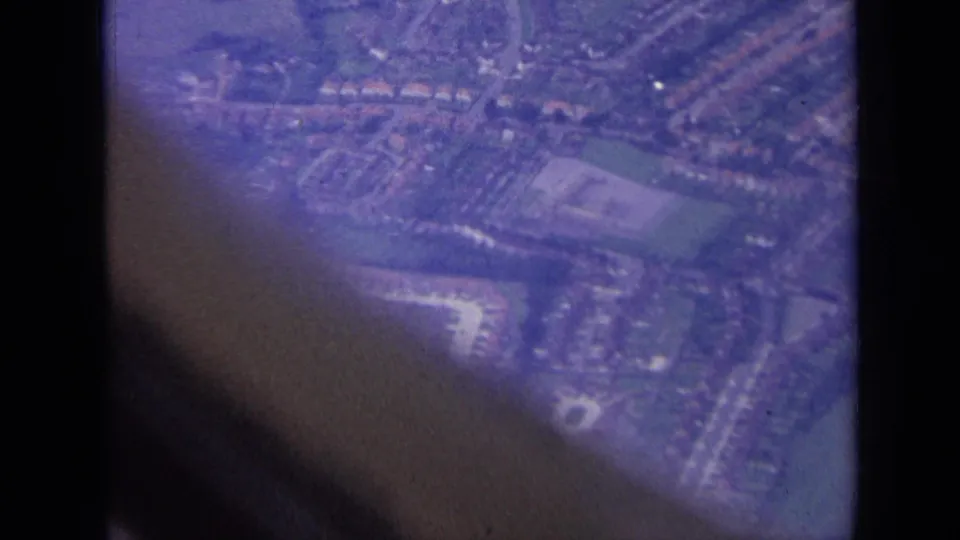 1963 aerial home-movie frame over London suburbs, with residential blocks, fields, roads, and dark aircraft-window obstruction.