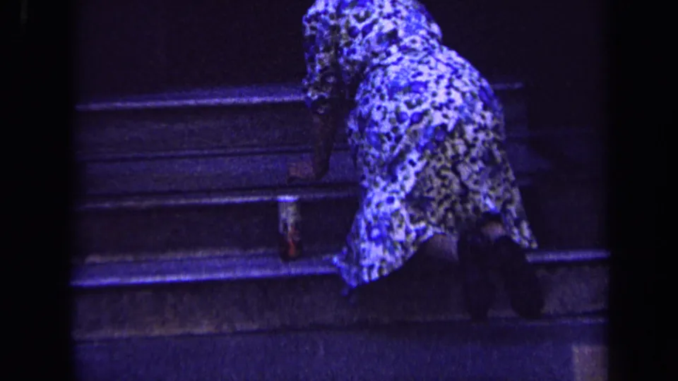 In 1963 London footage, a woman in patterned clothing kneels on stone steps under blue-tinted light while cleaning the surface.
