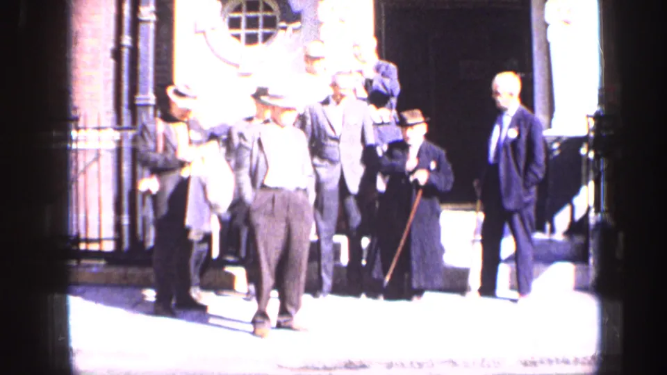 A group of formally dressed men stand outside a London building entrance in bright 1963 home-movie footage.