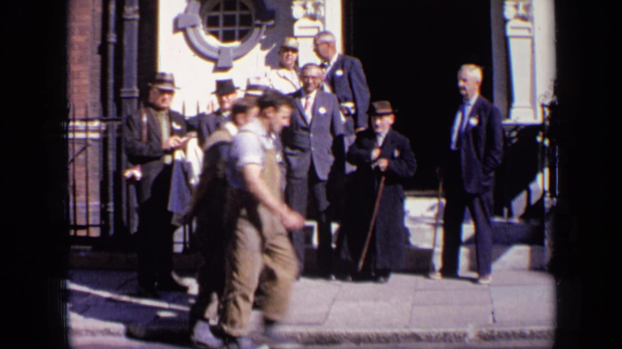 A passerby in work clothes crosses in front of suited men by a building.
