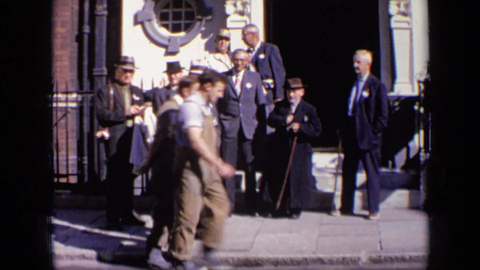 A man in work clothes walks through the foreground while formally dressed men remain outside a London building in 1963 footage.