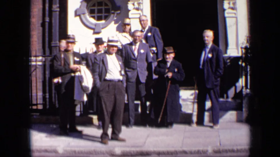 Several formally dressed men stand near a decorated building entrance in 1963 London, with strong sunlight and doorway shadow.
