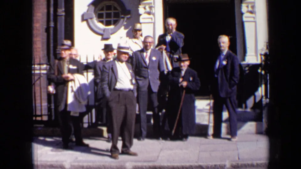 Men in mid-century suits and hats stand outside a London building in 1963, some facing the camera and one holding a cane.