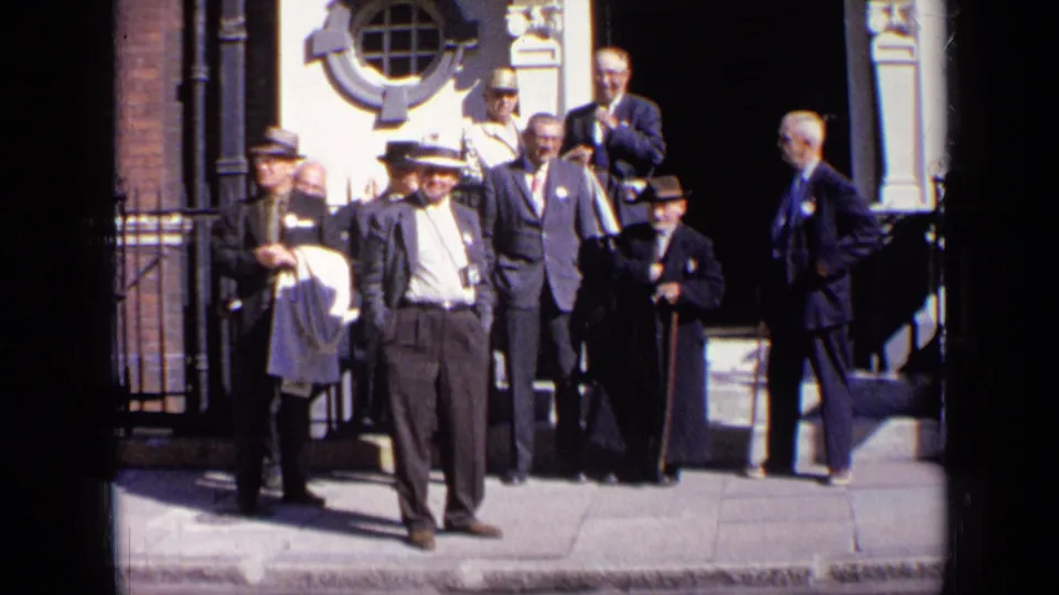 A formally dressed group of adult men remains gathered on a London sidewalk in 1963, framed by steps, railings and a circular window.
