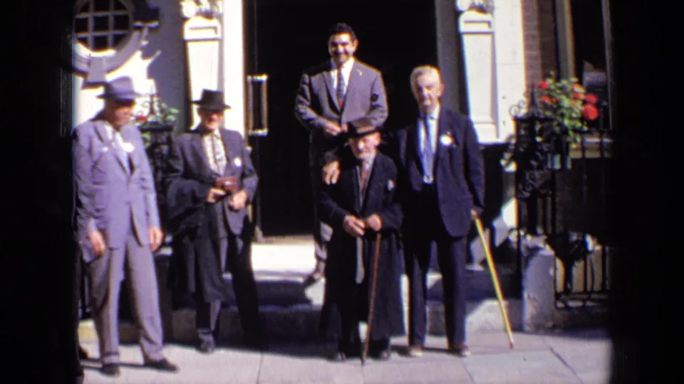 Five men in 1960s formal dress stand outside a London building, including an elderly man centered with a walking stick.