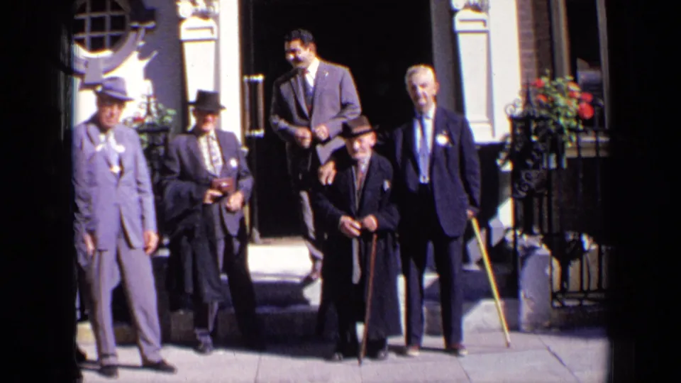 The 1963 London home-movie frame shows formally dressed men adjusting their stance outside a civic-looking entrance.