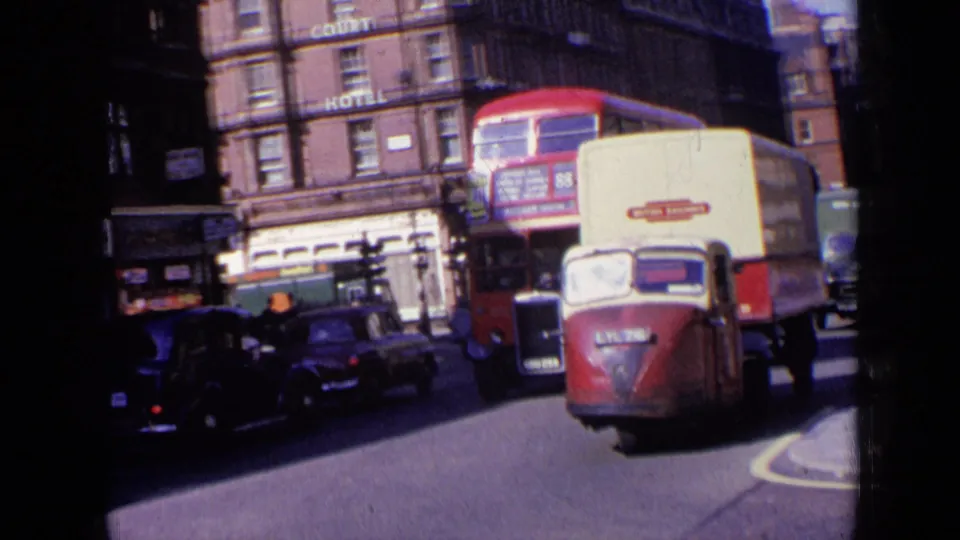 1963 London roadway with a red-and-cream delivery van in front, a red double-decker bus behind it, parked cars, pedestrians, and hotel signage.