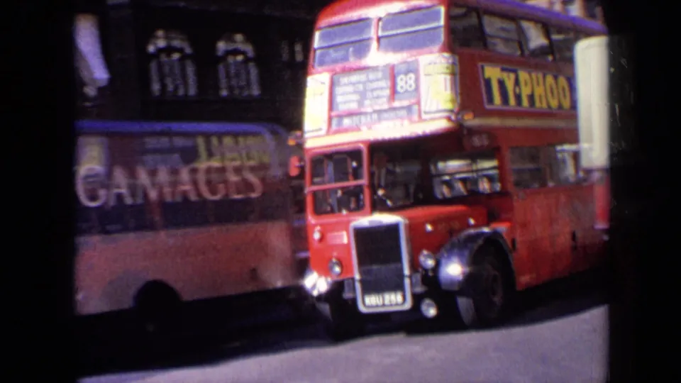 Close 1963 London street view of a red double-decker bus passing in front of another bus or vehicle with large side advertising, with dark cars nearby.