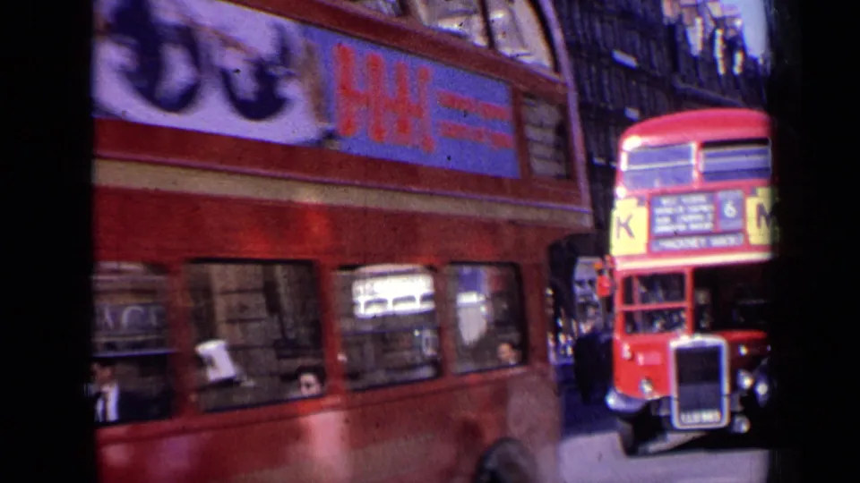 Two red London double-decker buses share the street, with advertising panels, pedestrians, and building fronts visible in the background.