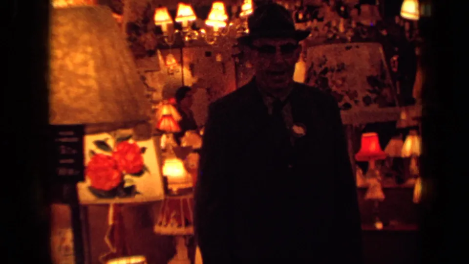 A man in a dark suit, tie, and brimmed hat stands in a warm London showroom surrounded by table lamps, chandeliers, signs, and framed display material.