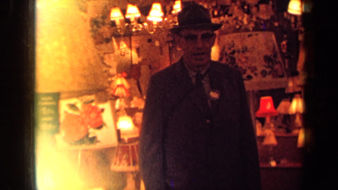 In 1963 London footage, a suited man in a fedora faces the camera amid glowing lamps and decorative shop displays.
