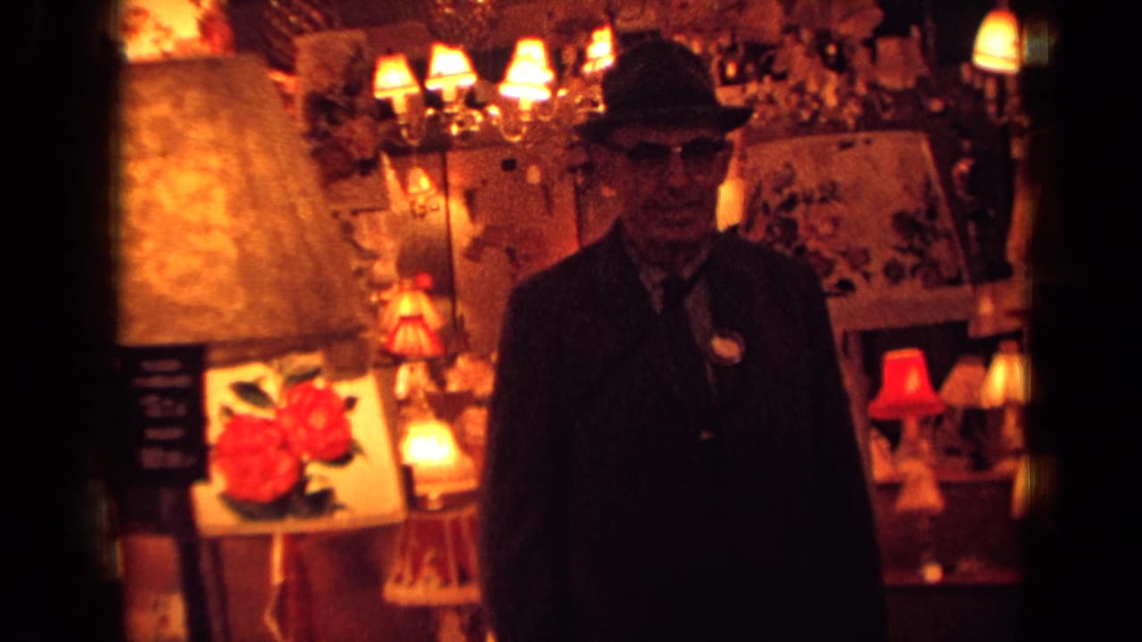 Man in dark clothing faces the camera beside bright lamps and wall displays.