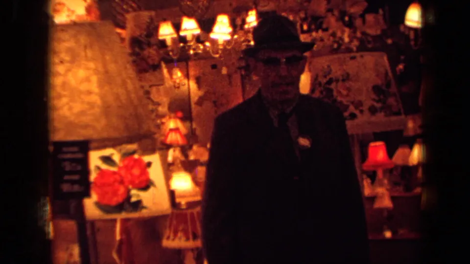 The 1963 London home-movie frame shows a suited man in a fedora near a large lampshade, small lamps, and red rose artwork.