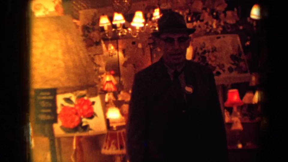A fedora-wearing man faces the camera in a warm, dim London shop interior with lamps, decor, and red rose artwork visible.