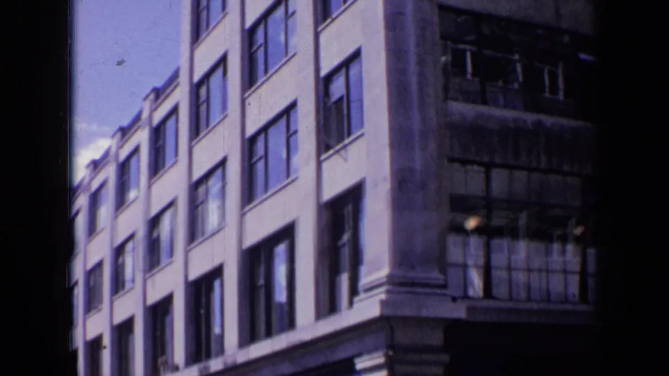 1963 London 8mm frame showing a pale multi-story building next to a darker weathered building with broken window panes.