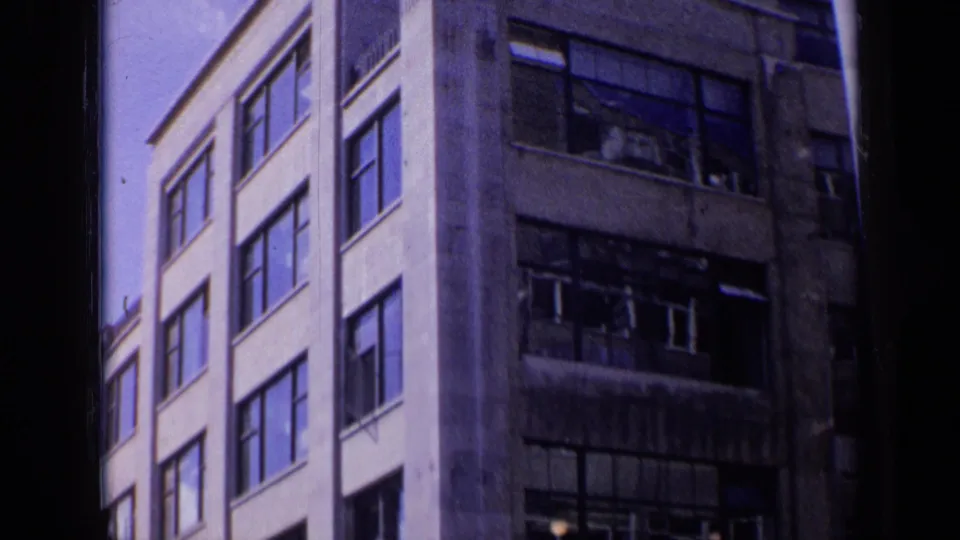 1963 London home-movie frame of adjacent multi-story buildings, one pale and one darker with visible broken glass.