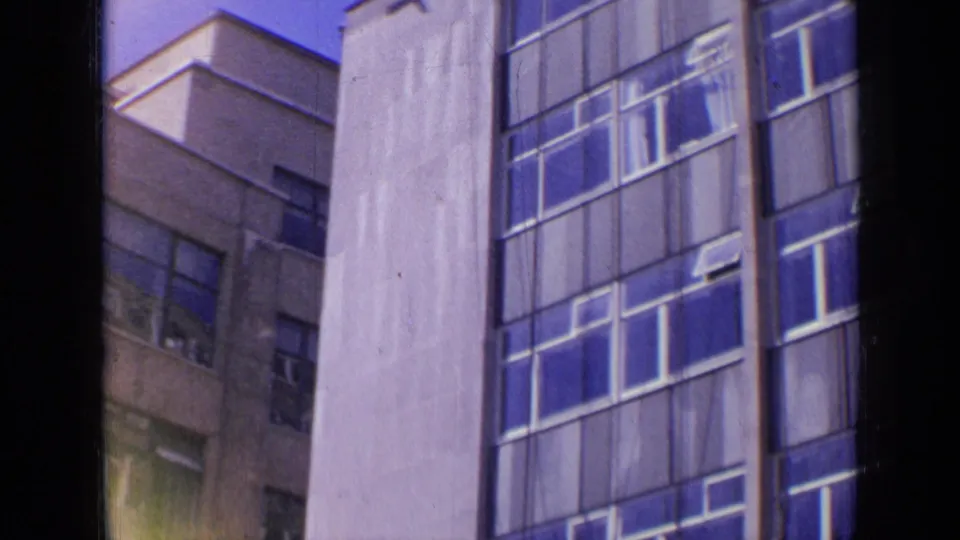 1963 London frame showing a pale multi-story wall with blue-tinted window grids and part of a darker building behind it.