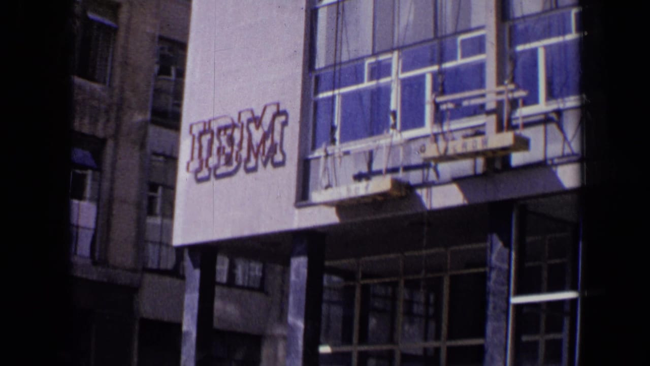 1963 London street view of a modern facade with IBM signage, large windows, deep shadows, and partial pedestrians at the edge of frame.