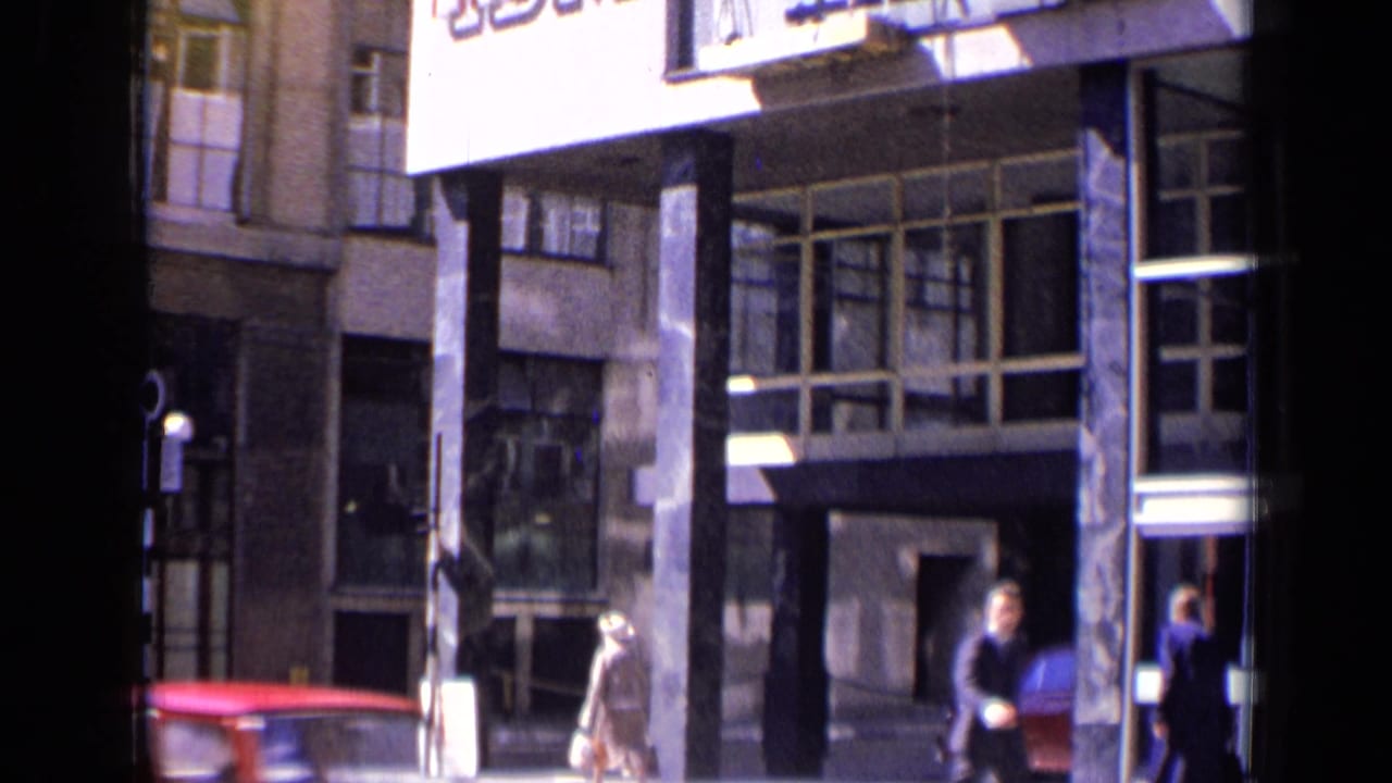 Street-level view of building columns, windows, pedestrians, and a red vehicle edge.