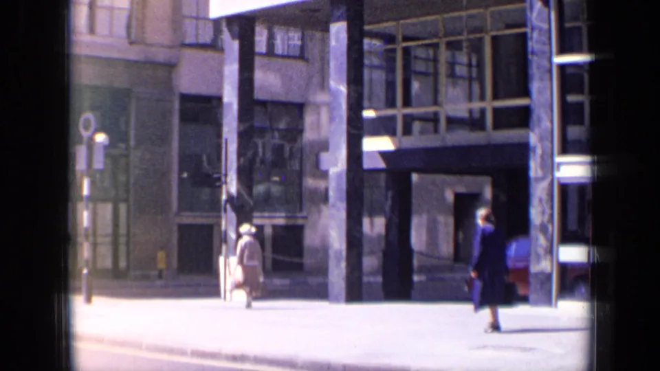 1963 London street view with a pedestrian walking past a modern building entrance, streetlamp, broad sidewalk, and a partial red vehicle.